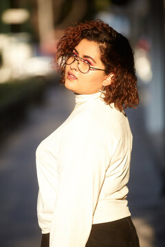 Side view of plump female wearing trendy clothes and glasses standing in city on sunny day and confidently looking at camera