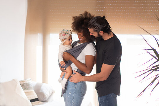 Side view of African American mother with toddler in baby carrier standing with ethnic father in cozy living room
