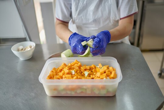 Female Cook In White Uniform And Protective Mask And Gloves Cutting Fresh Vegetables While Preparing Food In Hospital Kitchen During Coronavirus Pandemic