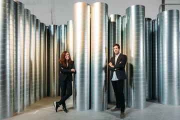 Smiling businesspeople in formal wear standing in contemporary industrial warehouse with metal barrels and looking at camera