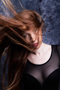 Calm female model in stylish clothes standing in studio with red flying hair and looking at camera