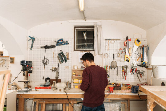 Back view of male woodworker standing at workbench and creating wooden details in bright garage