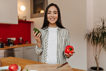 Joyful girl smiles, looking into camera in kitchen. Woman holding avocado and red pepper