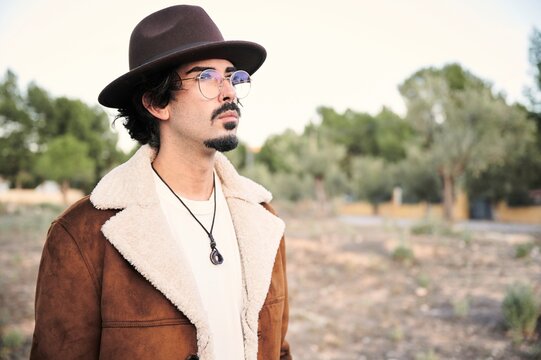 Handsome young bearded male in eyeglasses wearing trendy warm coat and hat looking away thoughtfully while standing against blurred trees in autumn day
