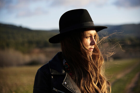 Stylish female wearing hat and black leather jacket standing in field at sunset and looking away