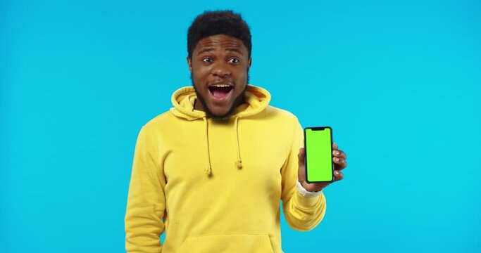 Close up portrait of handsome African American cheerful funny surprised young man holds in hand black smartphone with green screen isolated on blue background with impressed face, chroma key concept