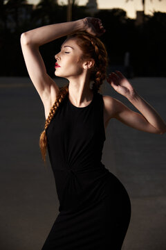 Slender stylish female model with braids and in black dress standing in street with eyes closed