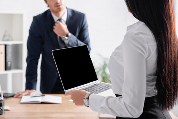 Fototapeta premium cropped view of sexy businesswoman holding laptop with blank screen near colleague adjusting tie on blurred background