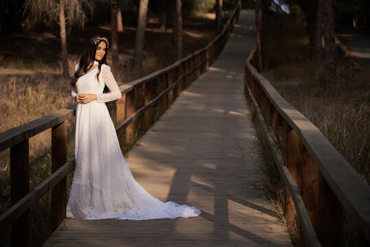 Full body side view of happy elegant young female in boho bridal maxi dress and wreath standing on wooden walkway in green woods and looking away