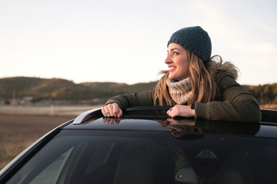 Cheerful female in warm clothes standing out of car sunroof and admiring amazing natural scenery during road trip