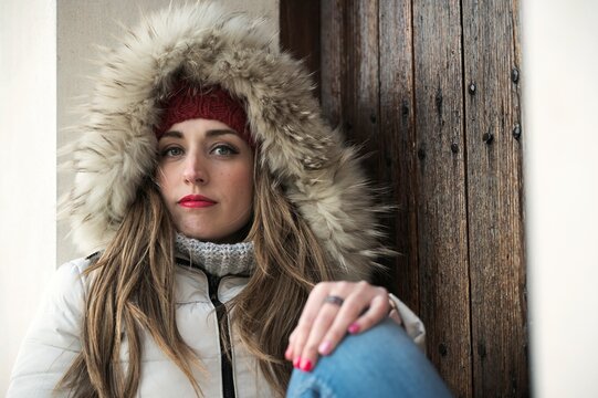 Tranquil female wearing warm winter jacket and knitted hat sitting near wooden door in city and looking at camera