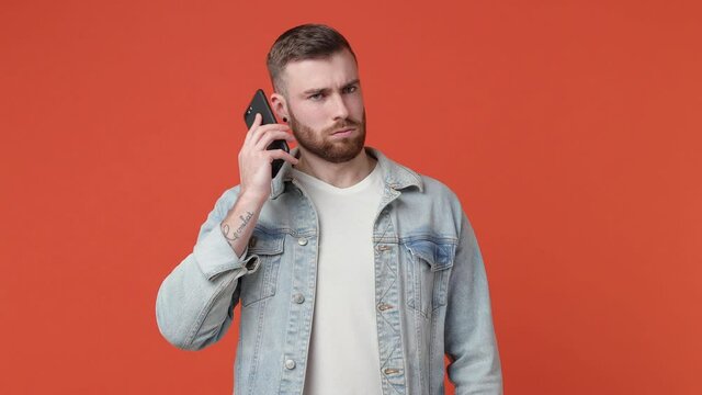 Irritated bearded young man 20s in casual white t-shirt denim jacket isolated on orange background studio. People lifestyle concept. Talk on mobile cell phone showing blah blah gesture ja jaja hands