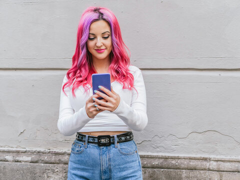 Modern alternative young female with long pink hair browsing on mobile phone while standing near stone wall on street