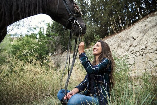 Tranquil female equestrian caressing muzzle of chestnut horse while sitting in woods and looking at each other