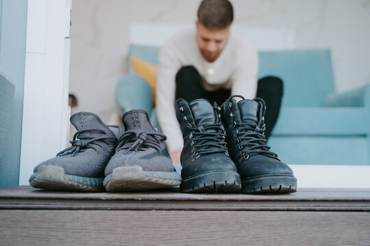 Ground level of dirty boots and sneakers placed at doorway of modern house of background of blurred man sitting on sofa