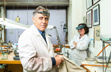 Side view of mature gray haired professional male jeweler in work wear and with glasses on forehead looking at camera while working with female colleague in workshop