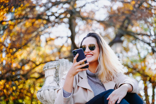 Low angle of cheerful female sitting in autumn park and texting on smartphone while enjoying conversation and looking away