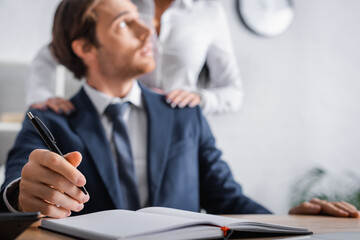businessman holding pen near notebook while secretary touching his shoulders, blurred background