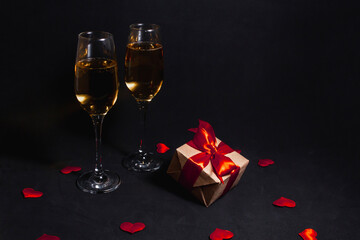 two glasses of champagne stand in the dark, next to a gift box tied with a red ribbon, and confetti made of hearts