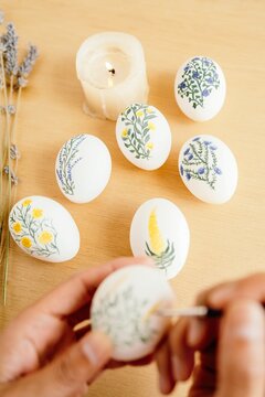 From above of crop unrecognizable person with paintbrush painting flowers on white eggs while preparing for Easter celebration at home