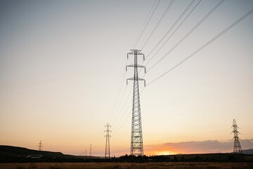 Low angle of tall electricity towers and wires in field under evening sky with sun setting over horizon line in summertime