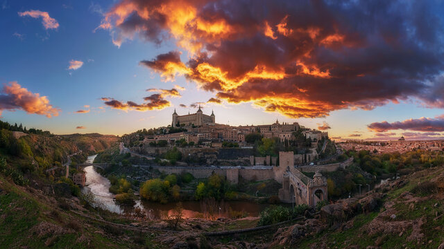 Panoramic view across river of old city Toledo in Spain with medieval castles and fortresses at sunset time with cloudy sky and reflection in river water