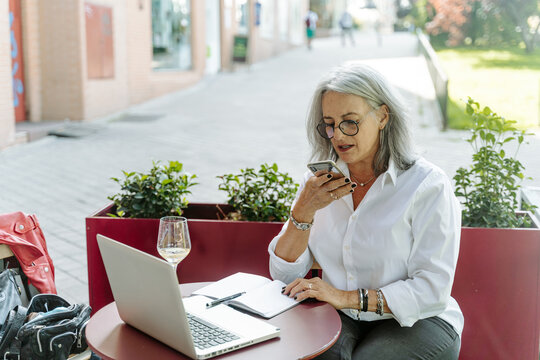 Busy middle aged female entrepreneur recording voice message on smartphone while working on project on terrace of cafe