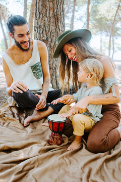 Delighted mother and father teaching little kid playing bongo drum while spending time together in woods