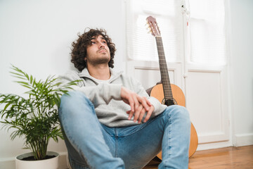 Young thoughtful hipster man in casual outfit sitting on floor near acoustic guitar and looking up while spending free time at home