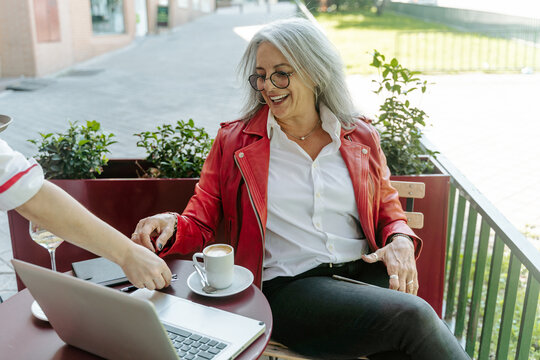 Crop waiter serving coffee for positive mature female entrepreneur sitting at table in outdoors cafe