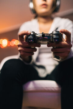 From below cropped blur unrecognizable concentrated female in headphones and with console sitting in armchair and playing video game while entertaining in evening at home