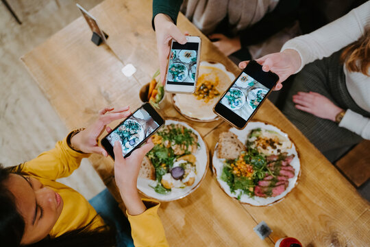 From above of diverse female friends gathering at table and taking photos of food on smartphones for social media