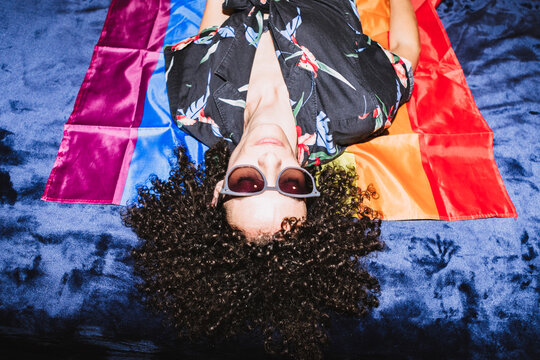 From above of calm upside down female in sunglasses lying on colorful LGBT flag in bedroom