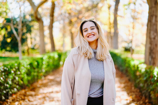 Smiling female wearing autumn coat standing in park on sunny day and looking at camera