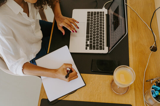 From above cropped unrecognizable female remote employee in casual outfit sitting at table with laptop and writing in planner while working in home office