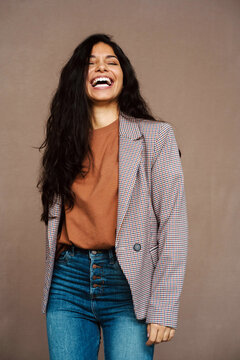 Delighted ethnic female in casual jacket laughing with closed eyes on brown background in studio