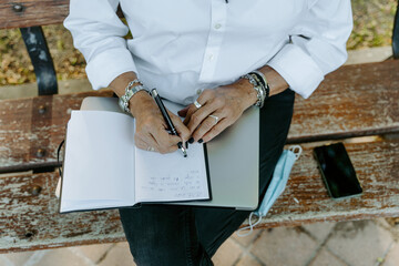 From above cropped unrecognizable female entrepreneur in smart casual wear sitting on bench and writing list of daily routine tasks while working remotely in park