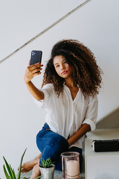Young ethnic curly haired female in casual white blouse and jeans taking self portrait on mobile phone while sitting on stairway at home