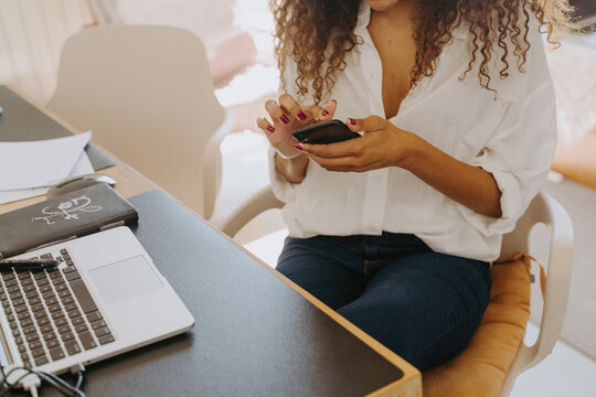 Cropped unrecognizable African American female entrepreneur with erabuds sitting at table in home office and reading messages on cellphone during distant work
