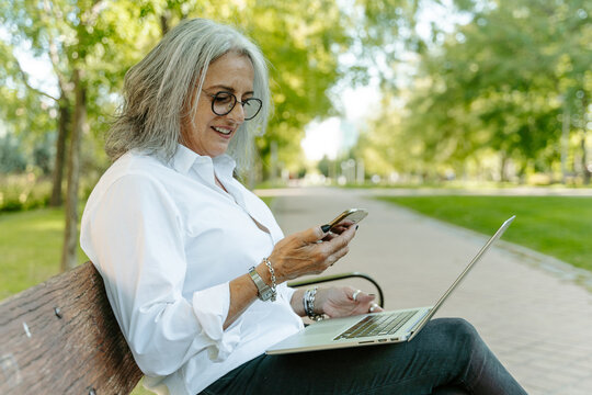 Side view of mature female entrepreneur sitting on bench in park and browsing smartphone while reading working messages