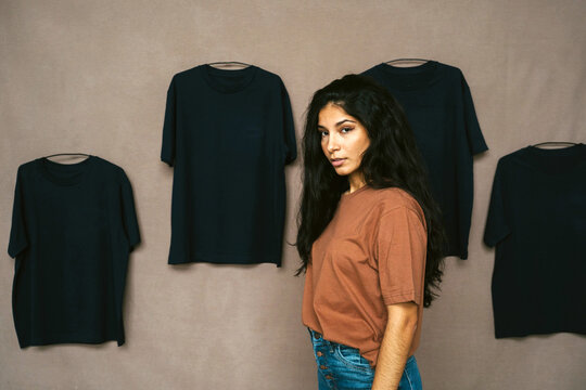 Side view of ethnic female in casual outfit standing in studio with t shirts on wall and looking at camera