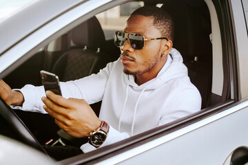 Side view of rich determined African American male driver sitting in parked car and browsing smartphone