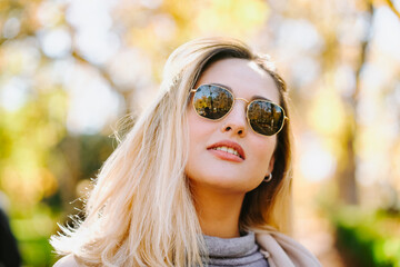 Smiling female wearing autumn coat and sunglasses standing in park on sunny day and looking at camera