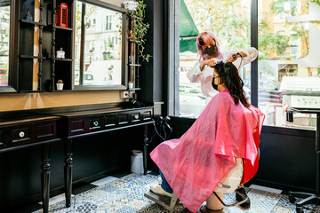 Side view of female hairdresser using curling iron and making trendy hairstyle for client in modern salon during coronavirus pandemic