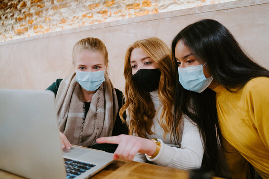 Group of multiethnic female freelancers in masks working on laptop in cafe while discussing new business project during coronavirus epidemic