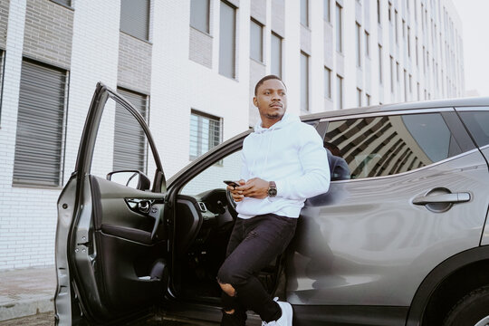 African American male standing near expensive car and surfing Internet on smartphone