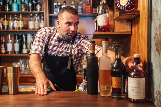 Crop brutal male bartender in apron grabbing bottle with alcoholic drinks placed on counter