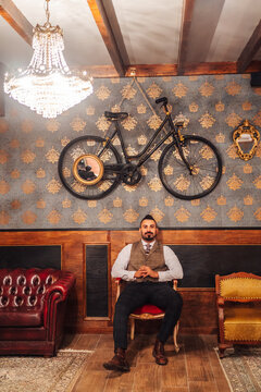 Full body of serious bearded well dressed man on classic suit sitting on chair in vintage restaurant