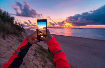 Back view of unrecognizable female traveler with long dark hair in casual clothes taking picture of amazing sunset over sea with sandy beach on smartphone during holidays in Cantabria