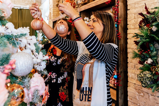 Side view of mature female friends buying christmas ornaments inside decor shop looking at baubles on christmas tree - Powered by Adobe